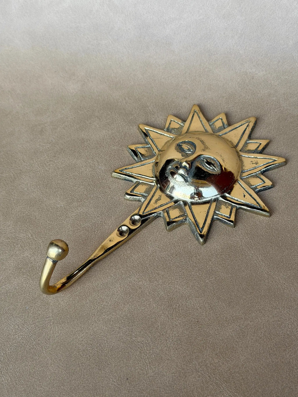 Brass sun face hook lying on its side on a taupe surface, showing the curved hook