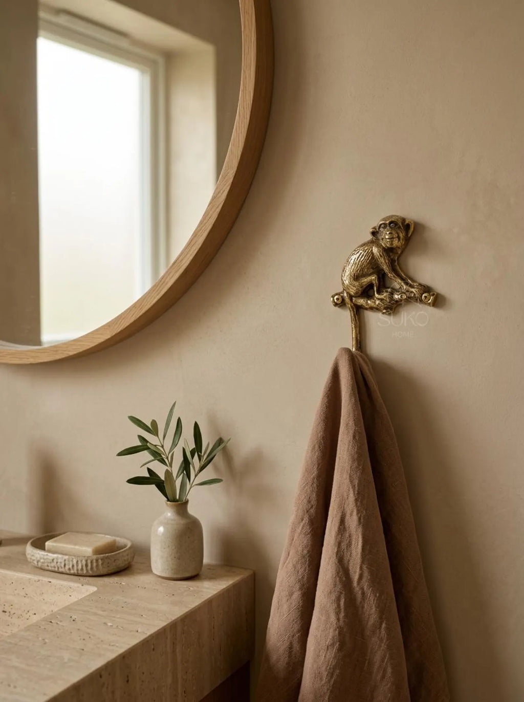 Brass monkey wall hook holding a brown linen towel on a beige wall beside a round mirror