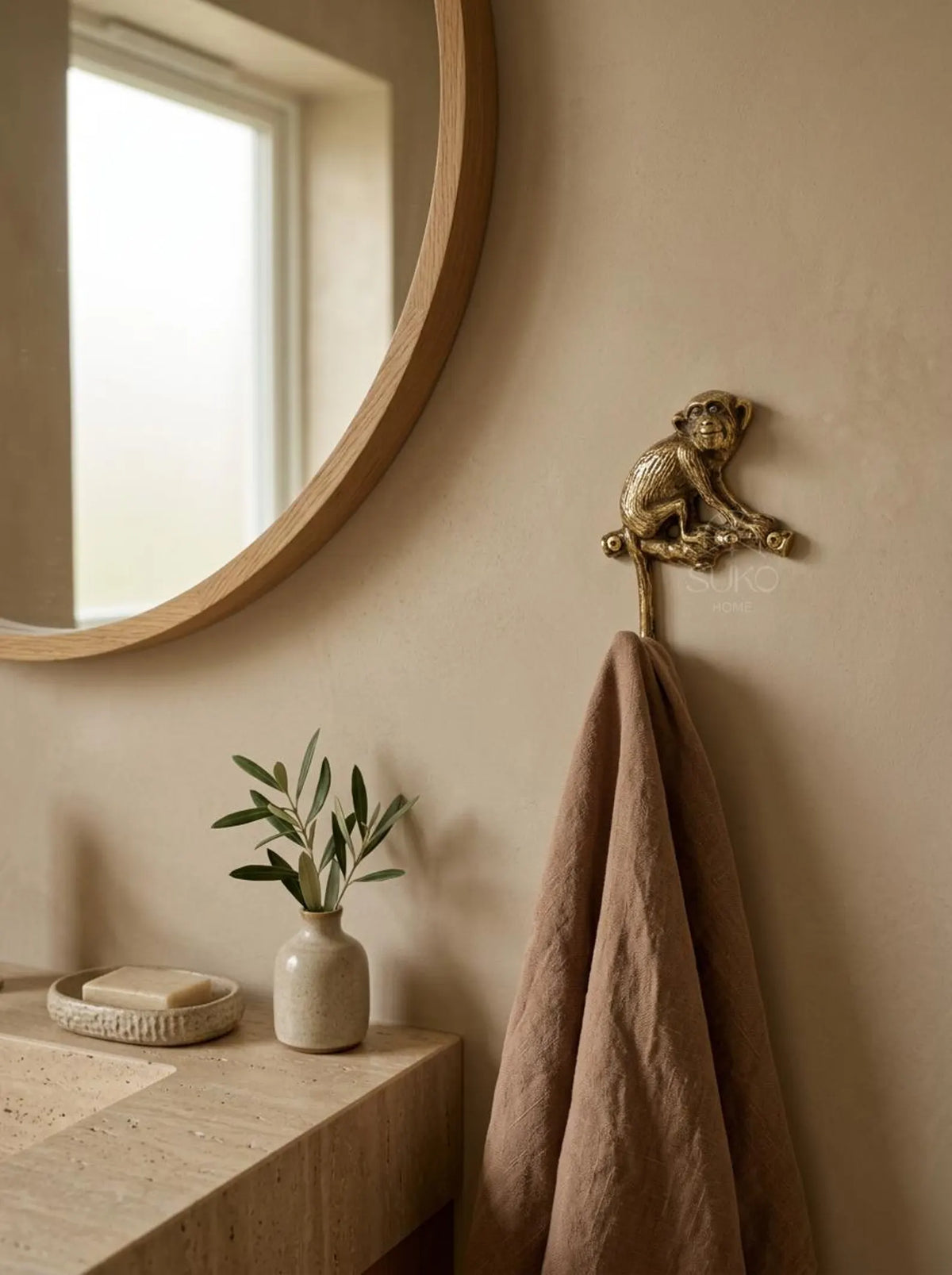 Brass monkey wall hook holding a brown linen towel on a beige wall beside a round mirror