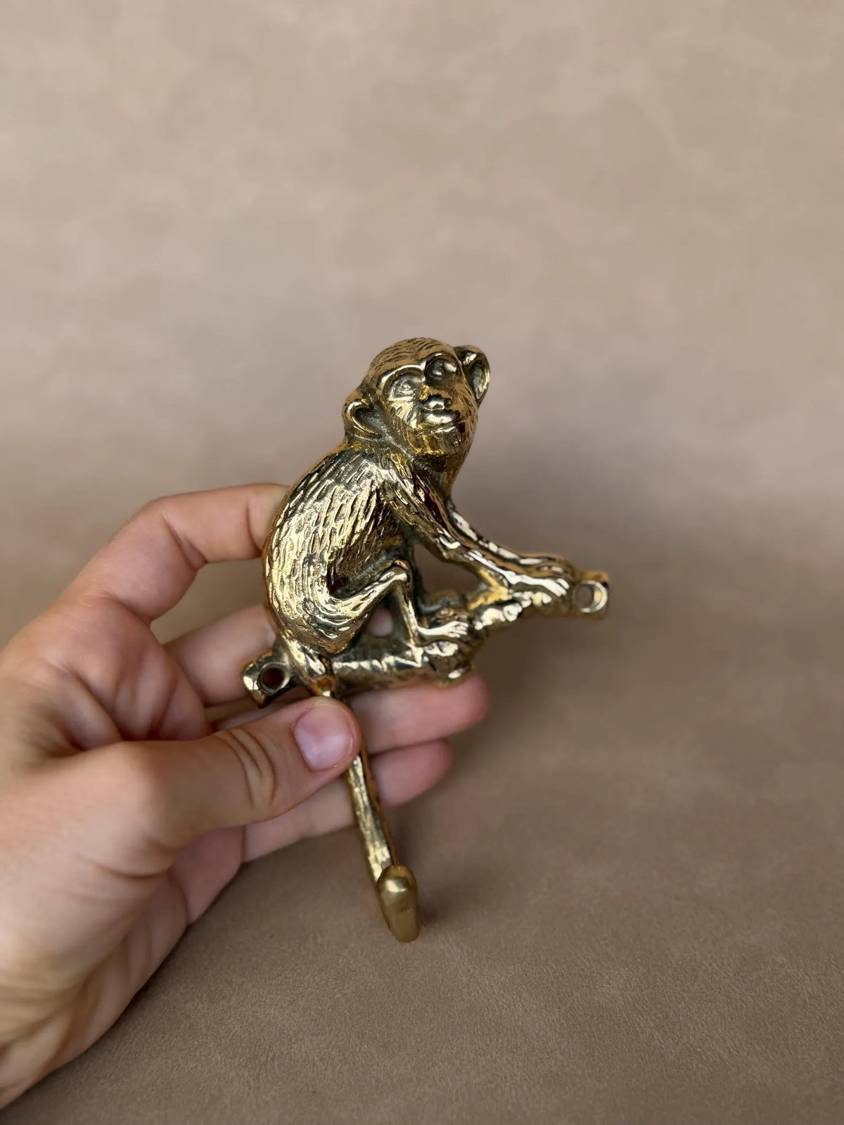 Hand holding the gold brass monkey hook at a slight angle against a taupe wall