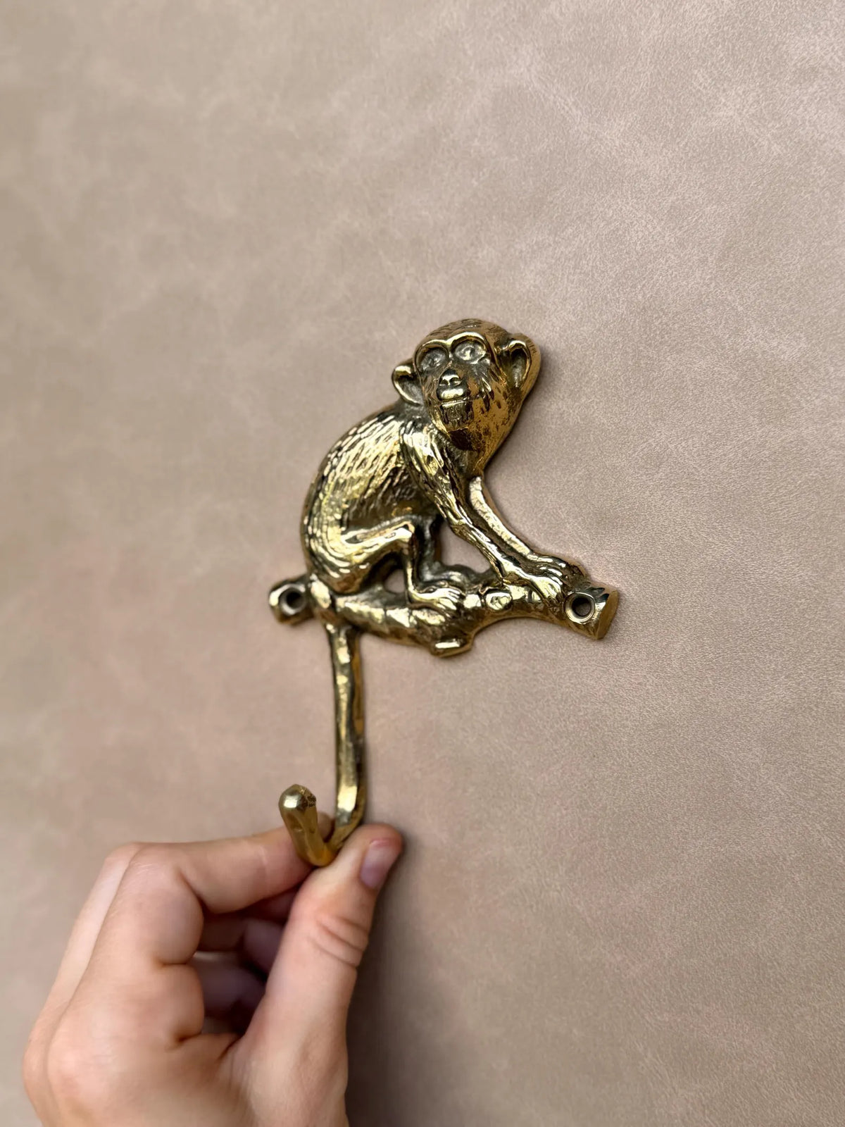 Hand holding the brass monkey hook upright by its curved hook, full length view