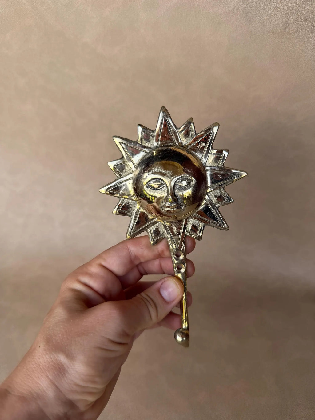Front view of the brass sun hook held by its hook, showing the calm human face