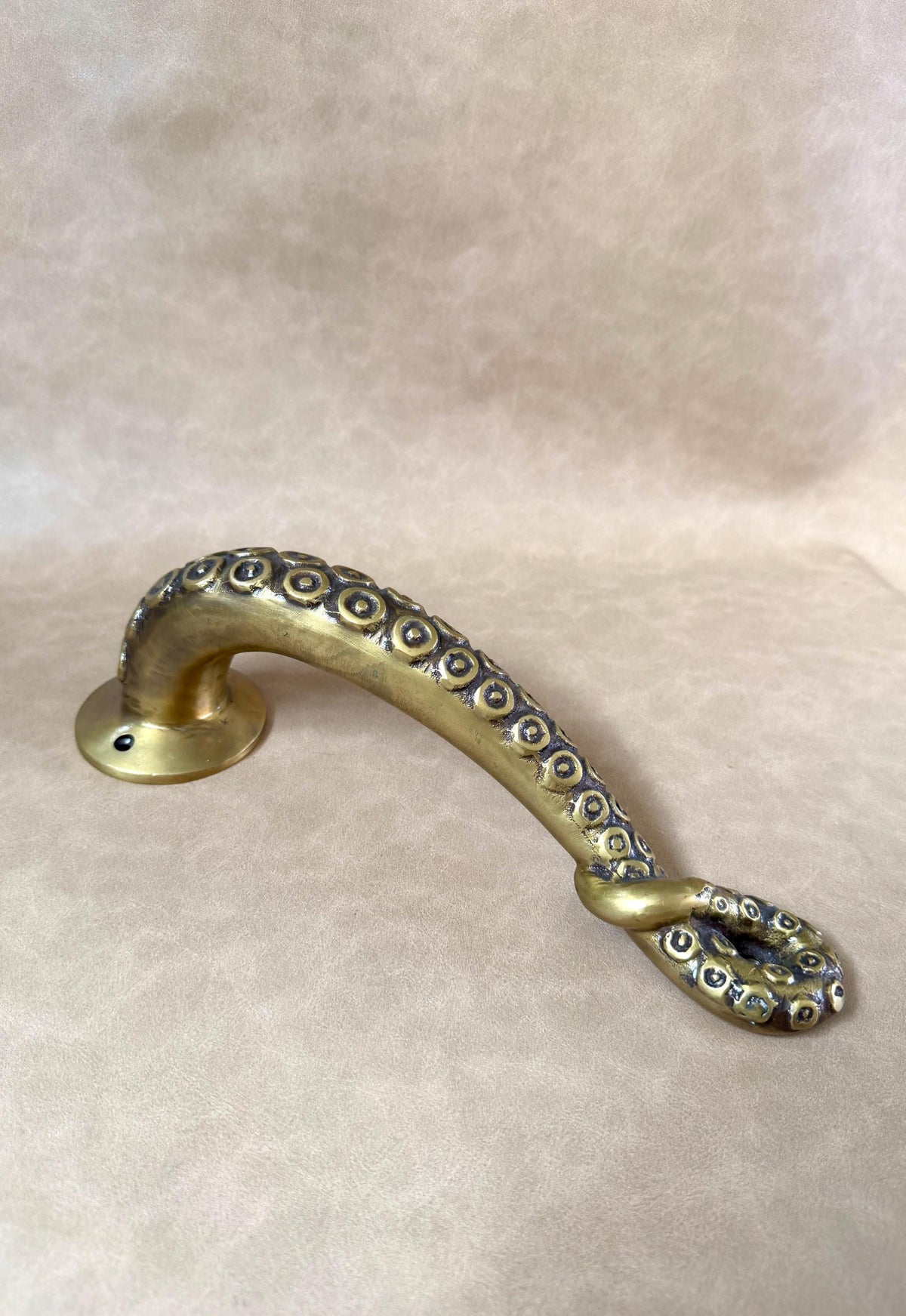 Brass octopus tentacle door handle lying flat on a beige background, showing its curve and mounting plate