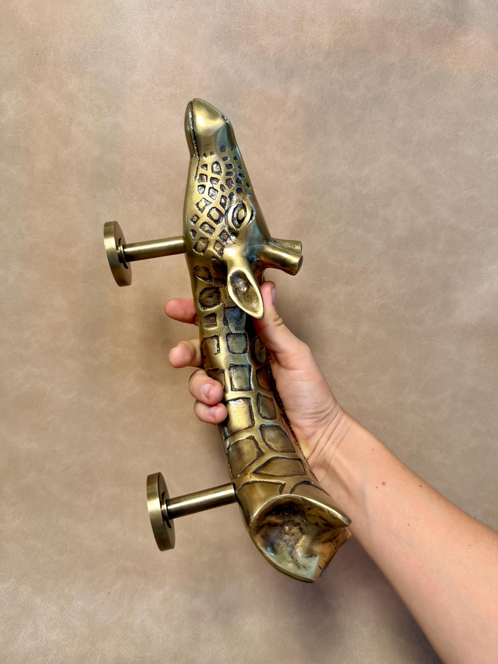 Hand holding the brass giraffe pull in an antique finish, with the patterned neck and two mounting posts visible