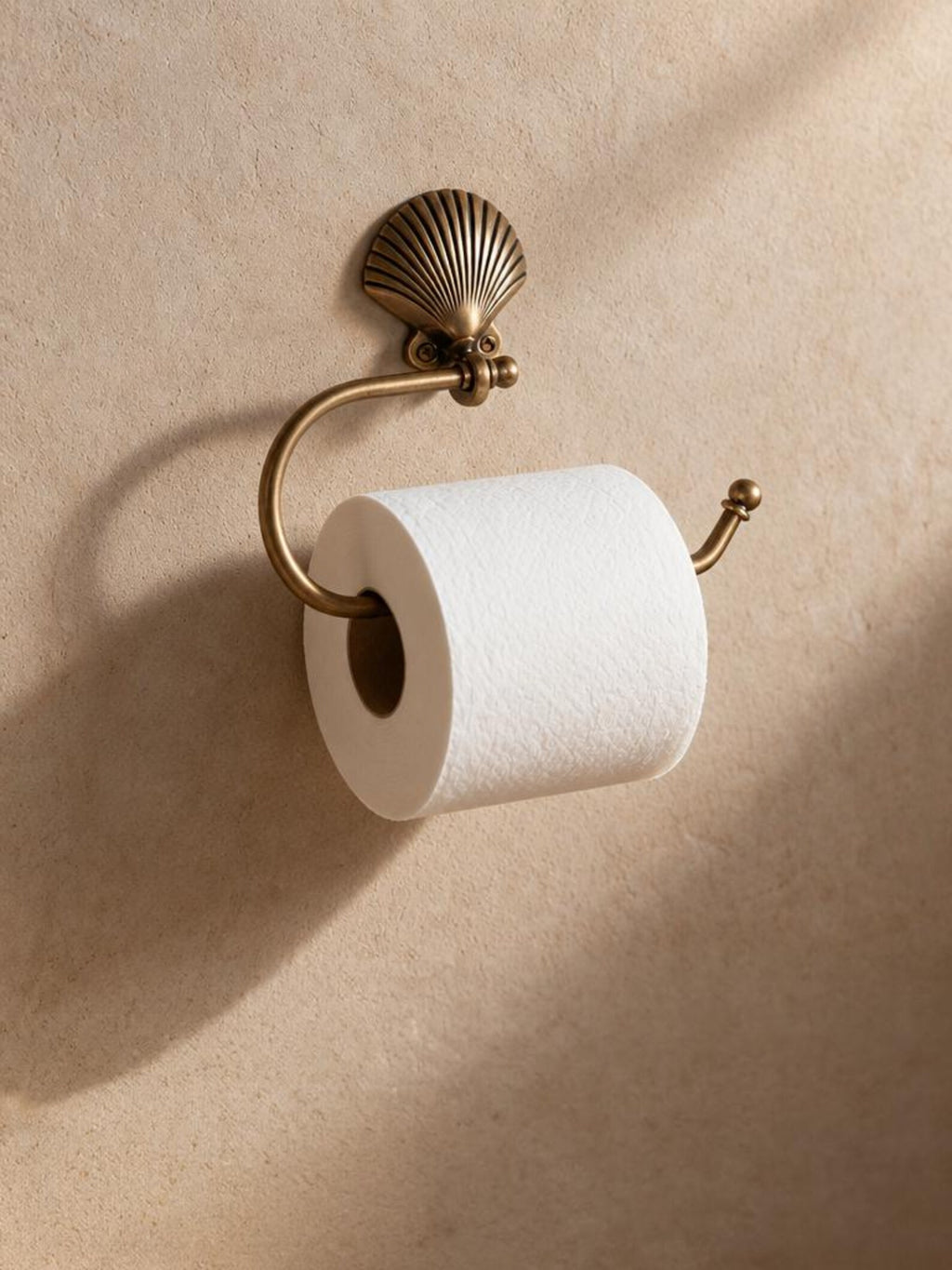 Brass shell toilet paper holder with a white roll on a warm beige wall
