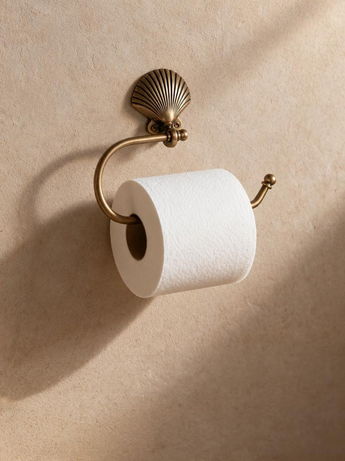 Brass shell toilet paper holder with a white roll on a warm beige wall