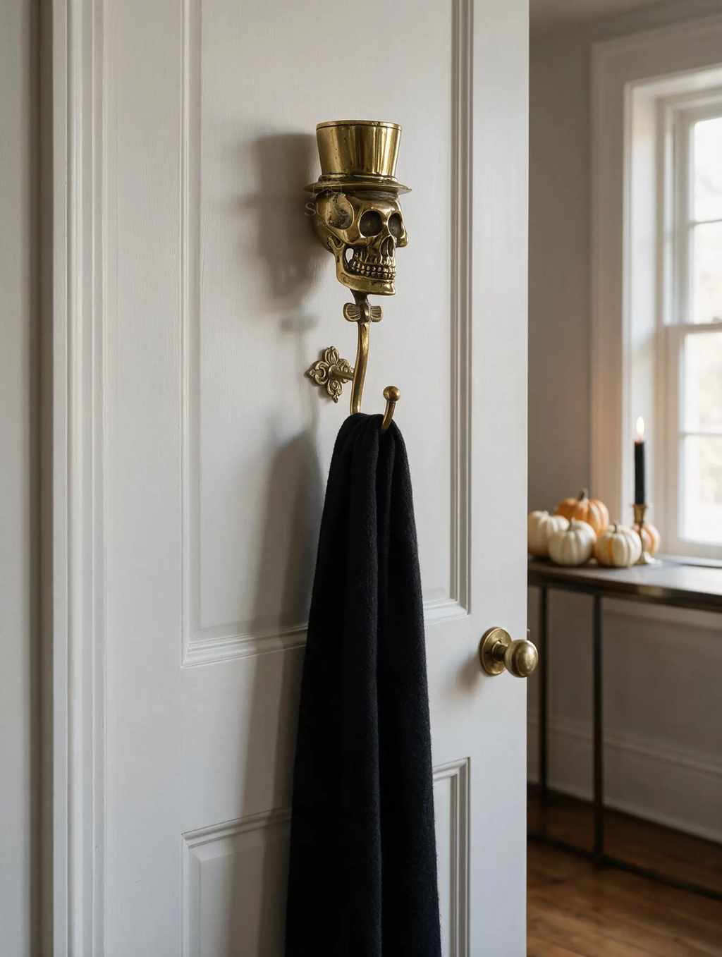 Brass skull wall hook with a top hat on a white door, holding a black towel, pumpkins in the background